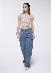 T-shirt cropped di colore rosa chiaro con una piccola grafica, abbinata a pantaloni cargo in denim blu dal taglio largo, dotati di molteplici tasche e vita alta.