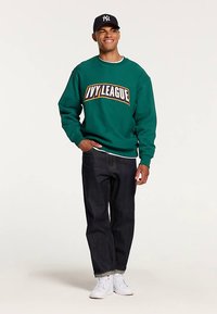 Vihreä collegepaita, jossa valkoisella ja kullalla teksti "IVY LEAGUE", yhdistetty tummiin farkkuihin ja valkoisiin lenkkareihin. Malli käyttää mustaa lippalakkia.