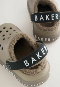 Zoccoli in schiuma marrone chiaro con cinturino nero contrassegnato "BAKER". Interno foderato in peluche, punta rotonda e fori di ventilazione lungo i lati.