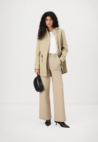 Giacca beige con cappuccio, top bianco e pantaloni beige a gamba larga. Borsa nera con una trama testurizzata e tacchi a punta neri.