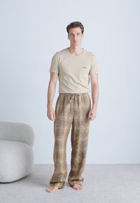 Beige cotton t-shirt with a small logo on the chest, paired with loose plaid pajama pants in brown and gold tones, featuring an elastic waistband.