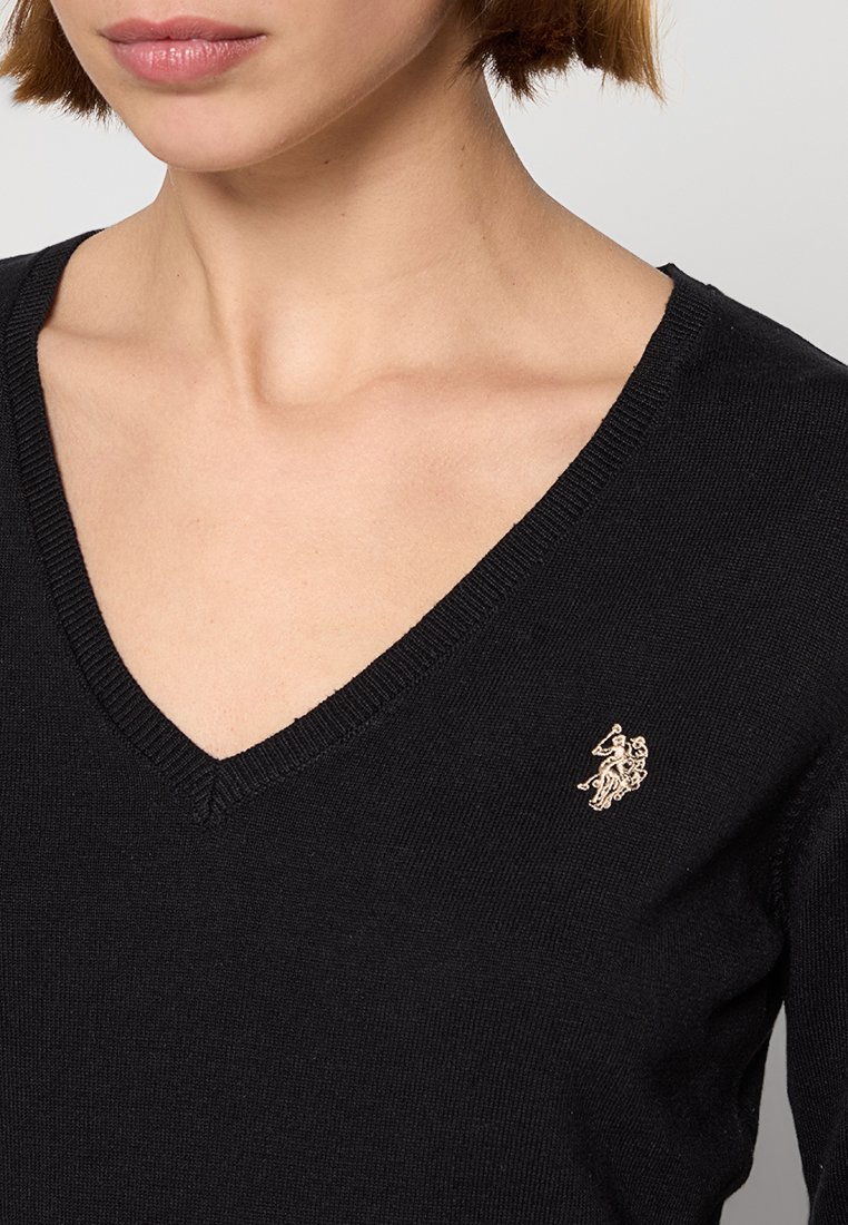 Pull en V noir en tissu tricoté doux avec un petit logo doré brodé sur la poitrine gauche. L'encolure présente des détails en côtes.