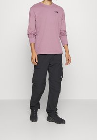 Dlouhý rukáv levandulové košile s černým logem, spárovaný s černými cargo kalhotami se zipovými kapsami a černými turistickými botami.