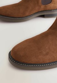 Botines de ante marrón con puntera redondeada, paneles laterales elásticos y costuras en contraste a lo largo de la suela. Textura suave y tacón bajo.
