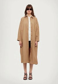 Világosbarna, túlnőtt ingruha, amely anyagból készült, gombos elülső résszel, zsebbel és laza fazonnal. Fehér ing és fekete nadrágshorts fölött viselve.