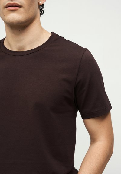 Νεαρό άτομο που φοράει ένα μονόχρωμο σκούρο καφέ κοντομάνικο μπλουζάκι, ορατό από τον λαιμό μέχρι τη μέση, με φωτεινό φόντο.