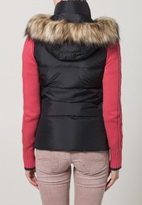 Svart vadderad väst med foder av pälshuva, i kombination med en rosa stickad tröja. Visar en figursydd design och kontrasterande texturer.