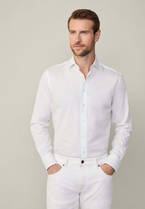 Camisa blanca de manga larga con botones, de textura suave y ligero brillo, combinada con unos vaqueros blancos. Presenta un cuello estándar y botones en la parte delantera.