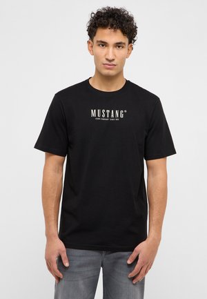 Giovane uomo con capelli ricci che indossa una maglietta nera con il marchio Mustang e jeans grigi, in piedi davanti a uno sfondo bianco uniforme.