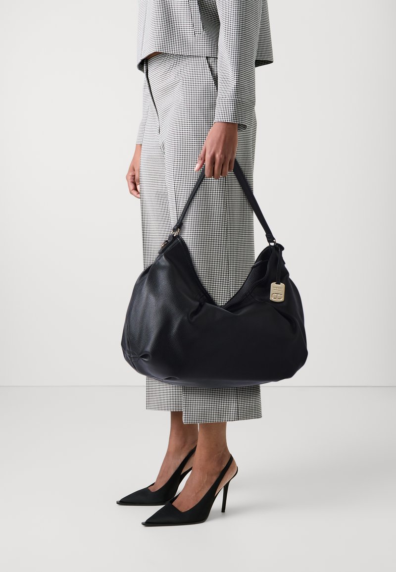 Femme portant un costume à carreaux noir et blanc, tenant un grand sac à main en cuir noir, chaussée de talons aiguilles slingback noirs sur fond blanc.