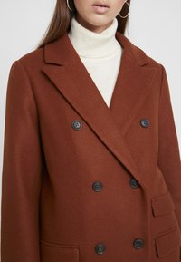 Brun uldblandings dobbeltbrystet frakke med klippet lapel, prydet med sorte knapper og en brystlomme, lagdelt over en cremefarvet rullekrave.