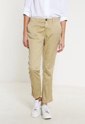 Persoon draagt beige rechte broek, opgerolde witte mouwen van een overhemd en witte sneakers, staand tegen een effen lichte achtergrond.