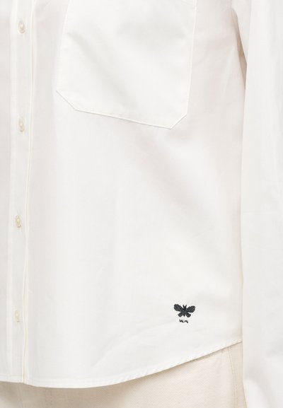 Chemise blanche boutonnée avec poche poitrine et petit logo papillon noir près de l'ourlet inférieur sur un pantalon de couleur claire.