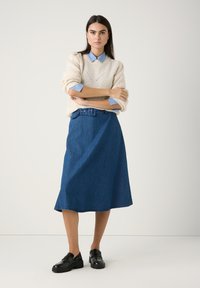 Strickpullover in Creme, hellblaues Hemd mit Kragen und einen dunkelblauen Jeansrock in A-Linie mit einem Taillengürtel. Schwarze Loafers runden das Outfit ab.