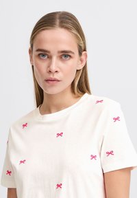 T-shirt en coton blanc à manches courtes avec des accents de nœuds roses brodés répartis de manière uniforme sur le tissu. Encolure ronde.