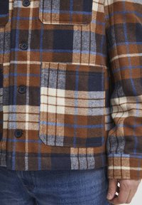 Brown and navy plaid flannel shirt with blue accents, featuring two chest pockets and black buttons. Soft texture, casual design.