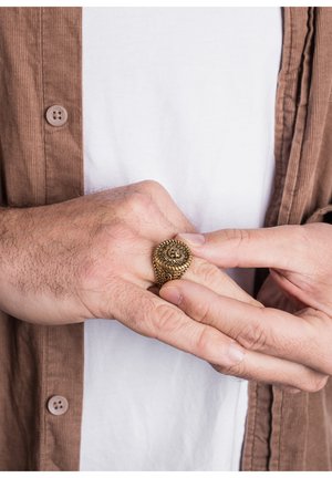 Inel cu model spiral din aur pe o mână, cu detalii intricate și design de filigran. Mâinile au o textură ușor vizibilă, pe un fundal neutru.