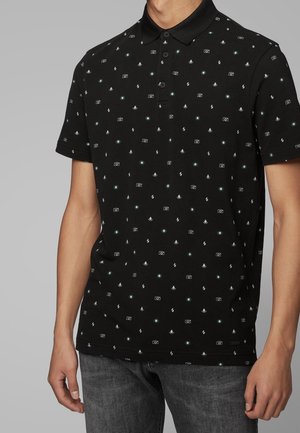 Mann trägt ein schwarzes Poloshirt mit kleinen weißen und grünen Symbolen von Kameras, Blitzzeichen und kreisförmigen Mustern, kombiniert mit dunkelgrauen Jeans.