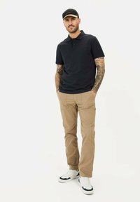 Marineblå polo t-shirt med to knapper, tanfarvede bukser og hvide sneakers. Har tatoveringer på armene og en sort kasket med røde detaljer.
