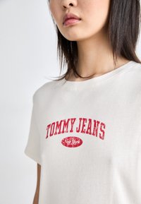Camiseta de ribete crema con mangas cortas y logo rojo "TOMMY JEANS" junto con el texto "New York", con un clásico cuello redondo.