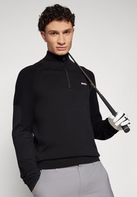 Črn zip-up golf pulover z rebrastim vzorcem na rokavih, s majhnim belim logotipom. Model drži golf palico in nosi rokavico.