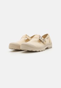 Zapatos slip-on beige con parte superior de malla, correa ajustable y suela de goma texturizada. Cuentan con una puntera redondeada y un tacón ligeramente elevado.