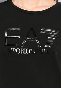 Černé tričko s velkým reflexním designem 'EAT' posetým kamínky a lesklým textem 'EMPORIO ARMANI' pod ním.