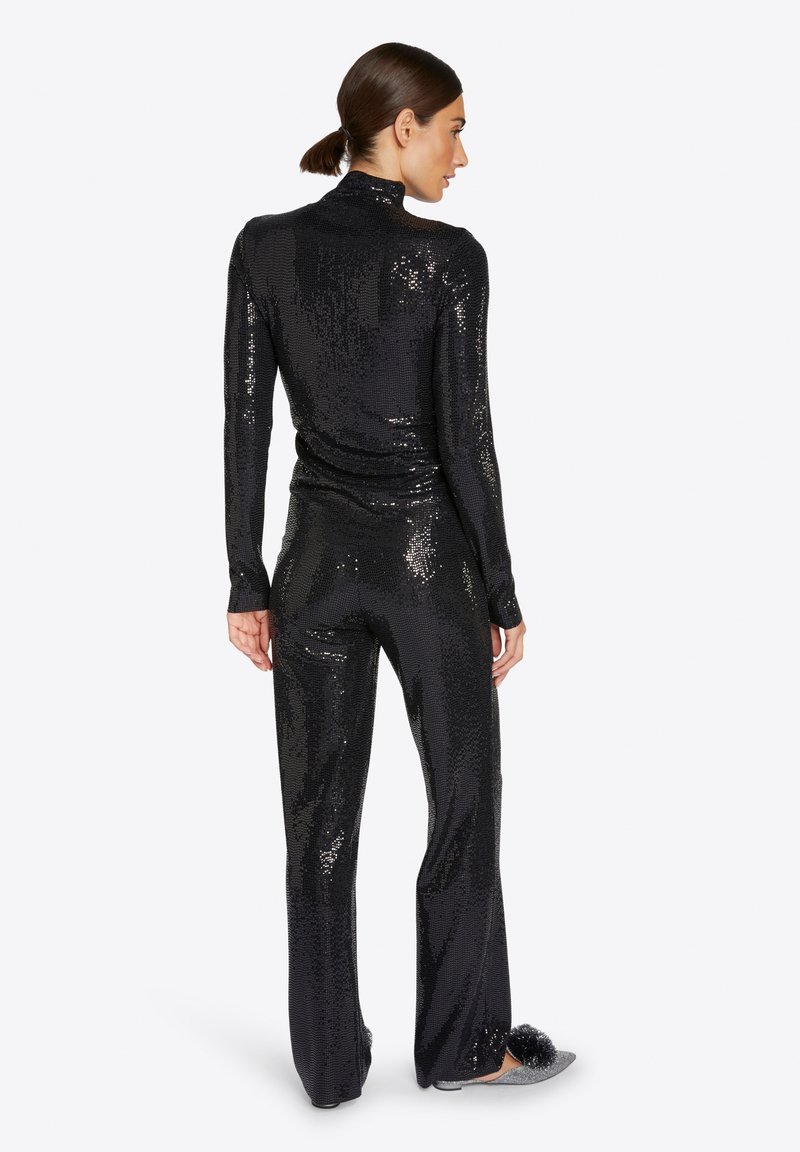 Kobieta odwrócona tyłem w czarnej, cekinowej bluzce z długim rękawem i dopasowanych szerokich spodniach, w srebrnych klapkach z czarnymi pomponami.