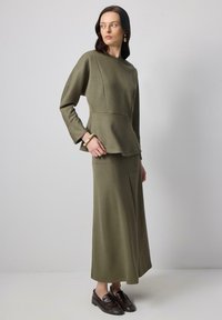 Alyvuogių žalios spalvos, ilgomis rankovėmis viršus su peplum dizainu, derinamas su atitinkančia ilga sijona. Glotni tekstūra, minimalūs siūlės, kartu su rudos spalvos loaferiais.