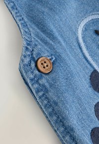 Acercamiento a una tela de mezclilla azul con costuras visibles, un botón marrón de cuatro agujeros de la marca "next" y un diseño bordado oscuro parcial.