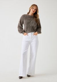Maglione marrone, morbido e a maniche lunghe, abbinato a jeans bianchi a vita alta e a gamba larga. Il modello sta in piedi con le mani nelle tasche, mostrando l'outfit.