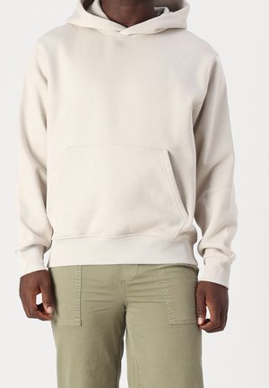 Hombre con sudadera con capucha de color beige claro con bolsillo canguro y pantalones verde oliva, de pie frente a un fondo liso.