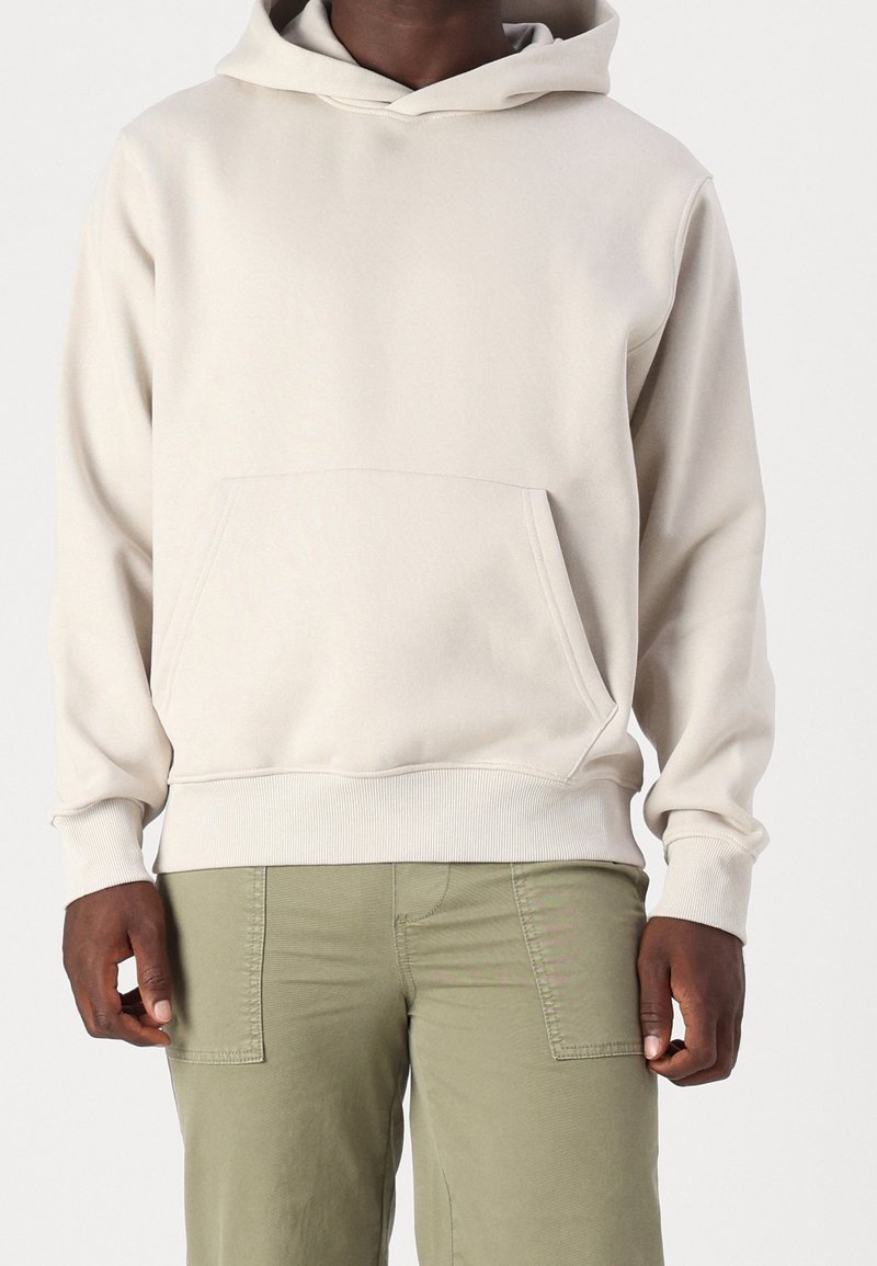Homme portant un sweat à capuche beige clair avec une poche kangourou et un pantalon vert olive, debout devant un fond uni.