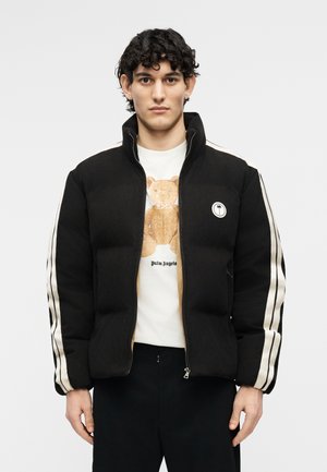 Νεαρός με σγουρά μαλλιά που φοράει μαύρο μπουφάν με άσπρες ρίγες στα μανίκια και λευκό t-shirt με γραφικό σχέδιο λούτρινου αρκουδάκι.