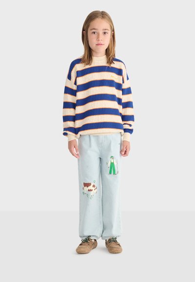 Enfant portant un pull rayé bleu et beige, un jean bleu clair avec des imprimés de fermier et de vache, ainsi que des chaussures marron, debout devant un fond uni.