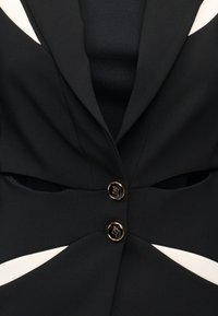 Μαύρο σακάκι ραμμένο με δύο χρυσά κουμπιά που φέρουν λογότυπο και λευκές τριγωνικές λεπτομέρειες κοπής στα πλαϊνά.