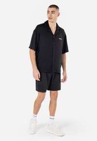 Chemise noire à manches courtes avec un col et des boutons, accompagnée d'un short en maille noir et de baskets blanches. Accentuée avec des chaussettes logo.