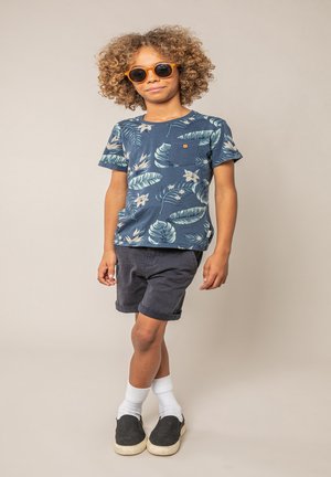 Enfant aux cheveux bouclés portant des lunettes de soleil orange, une chemise bleue à fleurs, un short sombre, des chaussettes blanches et des chaussures noires à enfiler, debout avec les jambes croisées.
