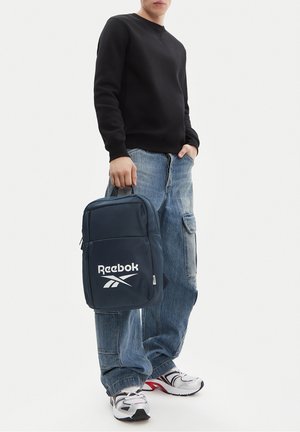 Osoba w czarnej bluzie i luźnych niebieskich dżinsach, trzymająca granatowy plecak Reebok w jednej ręce, stojąca w casualowych białych trampkach.