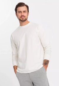 Camisa blanca de manga larga con escote redondo, hecha de una tela suave. El modelo tiene un tatuaje en la muñeca y lleva pantalones grises ajustados.