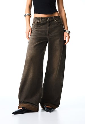 Personne portant un jean large marron foncé, un top court noir, des chaussures noires pointues et des bracelets noirs et blancs aux deux poignets.