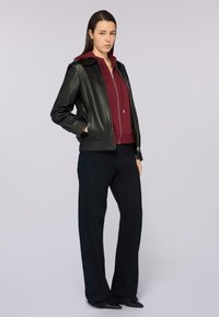 Chaqueta de cuero negra con cuello sobre una sudadera con cremallera color burdeos, combinada con pantalones oscuros de pierna ancha y zapatos negros puntiagudos. Texturas suaves.