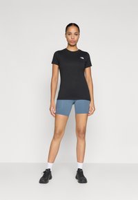 Melnas īsām piedurknēm t-krekls ar logotipu, savienots ar ziliem sporta šortiem un melnām apaviem. Fons ir vienkāršs balts.