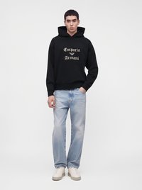Млад мъж, облечен с черен суичър Emporio Armani, светлосини дънки и бели маратонки, стои спокойно с една ръка в джоба.