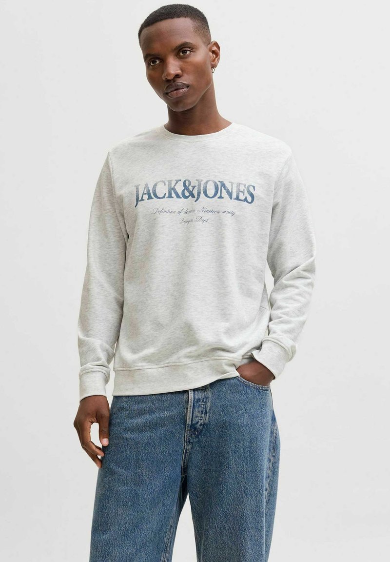 Sweat-shirt gris clair à manches longues, avec le texte bleu marine "JACK&JONES" et une impression supplémentaire. Porté avec un jean bleu. Design simple et décontracté.