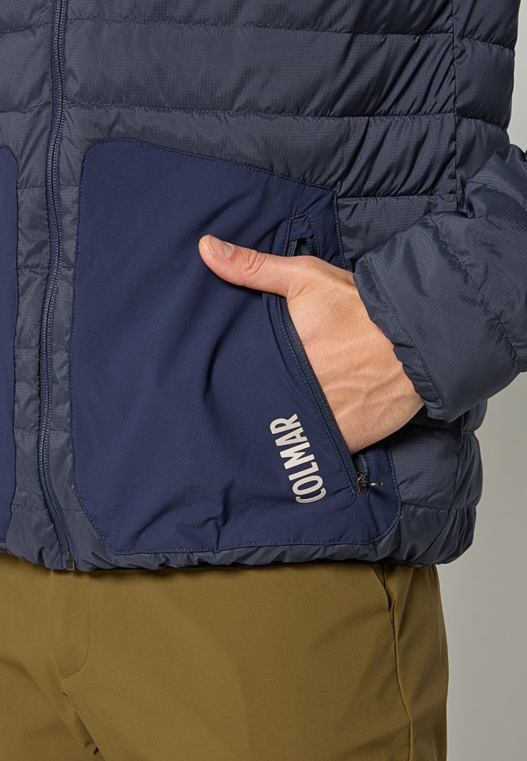 Veste matelassée bleu marine avec des coutures horizontales, poches zippées et détails de logo. Elle présente un tissu lisse et léger ainsi que des poignets élastiques.