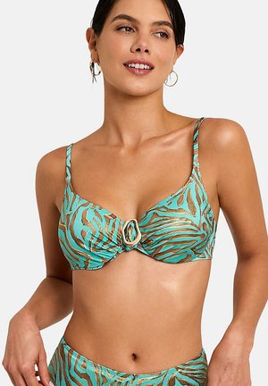 Vrouw draagt een turquoise en goud zebraprint bikini top met een centraal ringdetail, glimlachend met haar naar achteren en gouden oorringen.