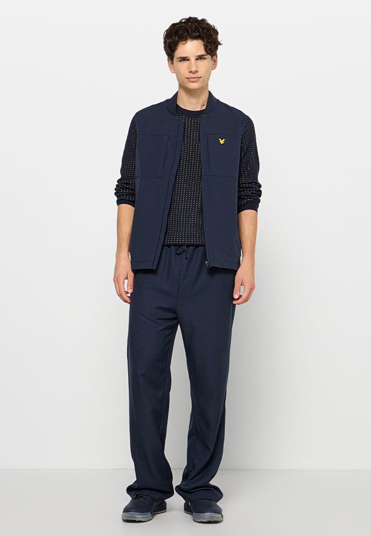Gilet zippé bleu marine avec poches avant sur une chemise noire à manches longues à motifs, associé à un pantalon bleu marine assorti et des chaussures sombres.