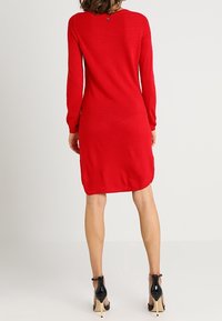 Robe rouge en tricot avec manches longues, col rond et détail de boutons au dos. Portée avec des chaussures à talons noirs.