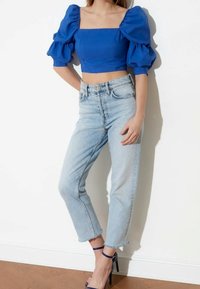 Kvinna som bär en blå puffärmad croptop, ljusblå högmidjade jeans och marinblå högklackade skor med ankelrem, stående mot en vit vägg.